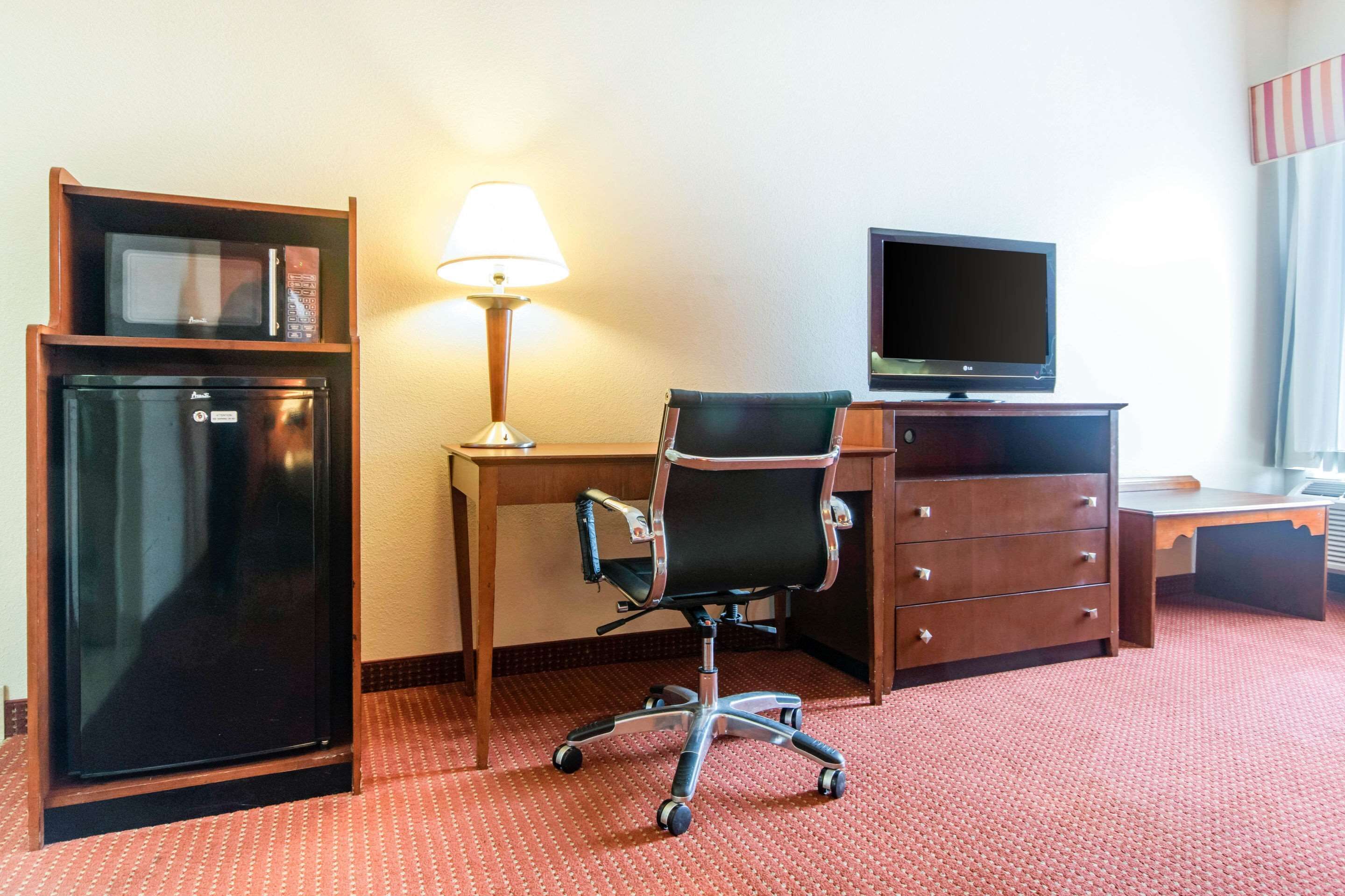 A hotel room with a desk, a chair, a television, a refrigerator, and a lamp.