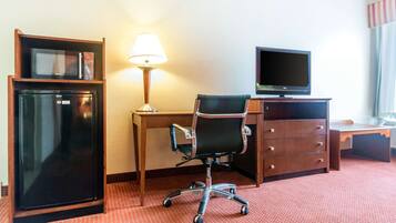 A hotel room with a desk, a chair, a television, a refrigerator, and a lamp.