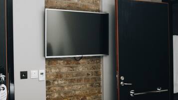 A bedroom with a brick accent wall, a flat-screen TV mounted above it, a door with a handle, and a light fixture with exposed bulbs.