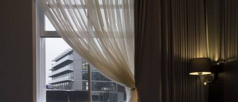 Hotelkamer met een bed, een raam met dunne gordijnen, uitzicht op een gebouw en een wandlamp.