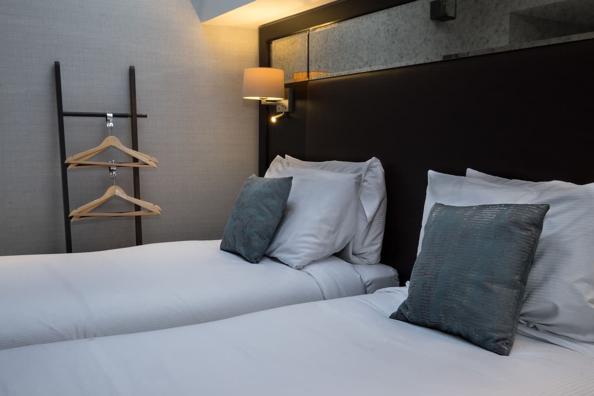 Hotellihuone, jossa on sänky, tyynyjä, vaateteline ja seinään kiinnitetty lamppu.