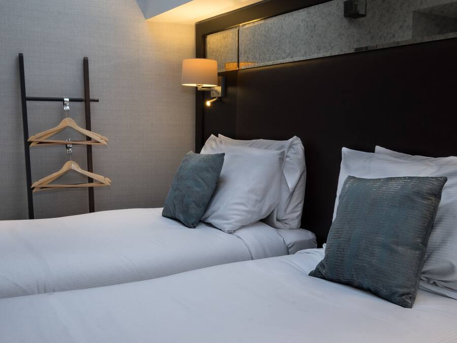 Habitación de hotel con cama, almohadas, perchero y lámpara fijada a la pared.