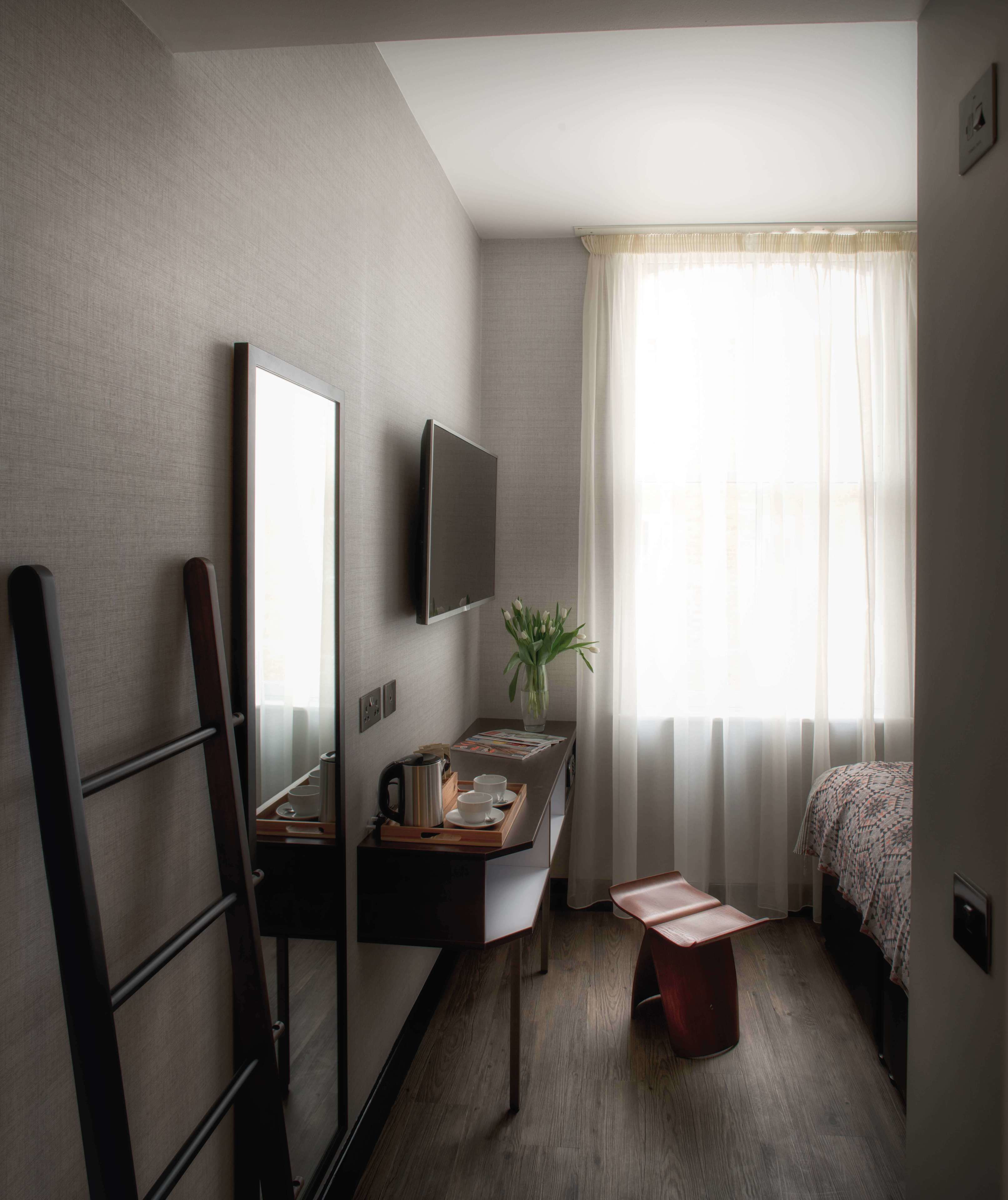 Quarto de hotel compacto com cama, escrivaninha com TV de tela plana, pequena mesa com um vaso de flores e janela com cortinas translúcidas.