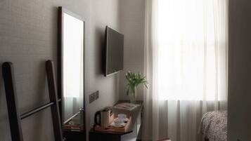 Quarto de hotel compacto com cama, escrivaninha com TV de tela plana, pequena mesa com um vaso de flores e janela com cortinas translúcidas.