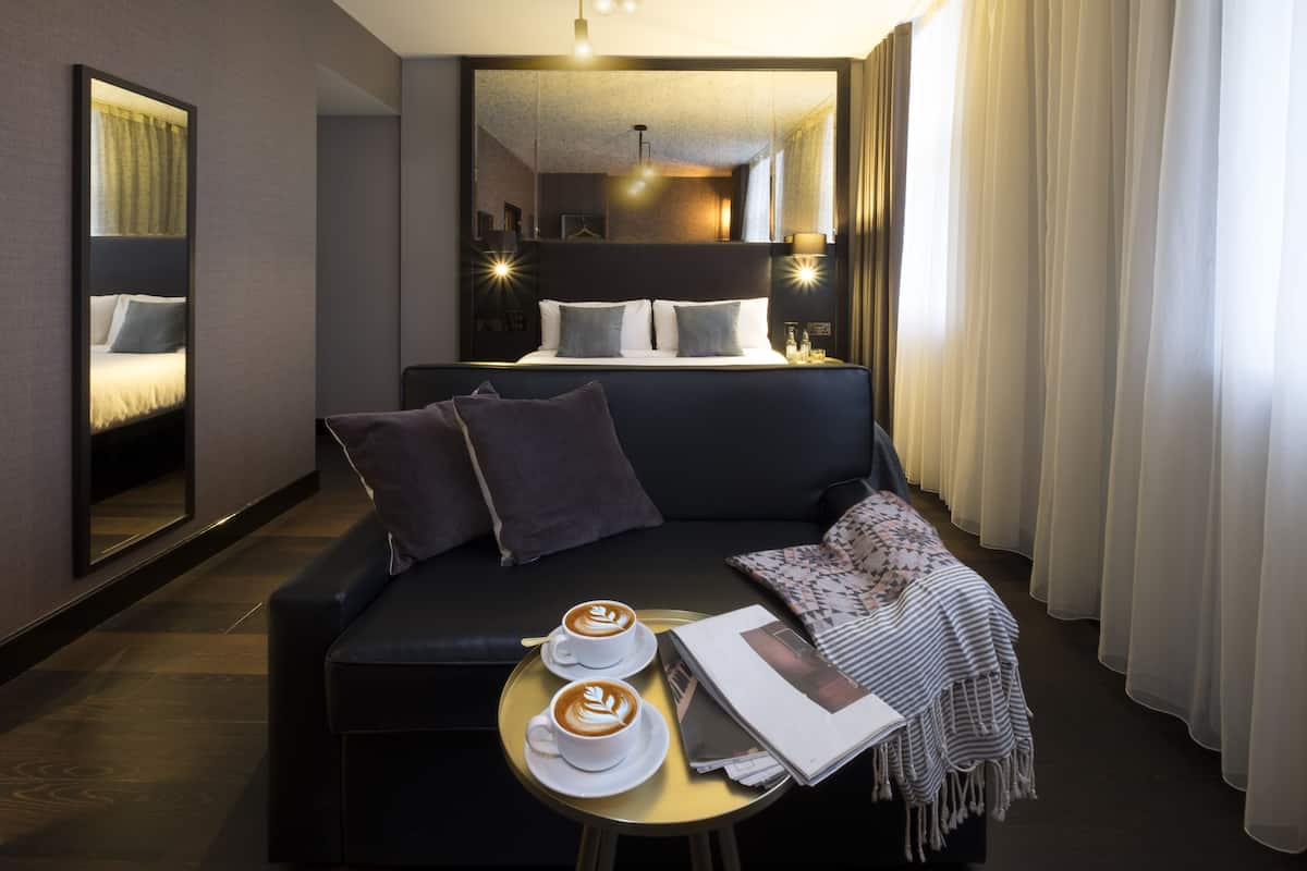 Moderni hotellihuone, jossa on sohva, pieni pyöreä pöytä, kaksi kahvikuppia, lehti ja sängynpääty.