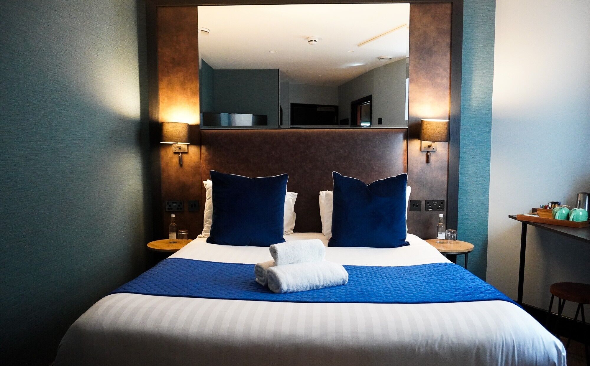 Habitación de hotel con una cama grande, dos lámparas de noche, un espejo sobre la cama y una mesita con una silla.