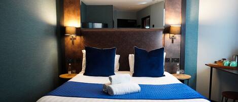 Habitación de hotel con una cama grande, dos lámparas de noche, un espejo sobre la cama y una mesita con una silla.