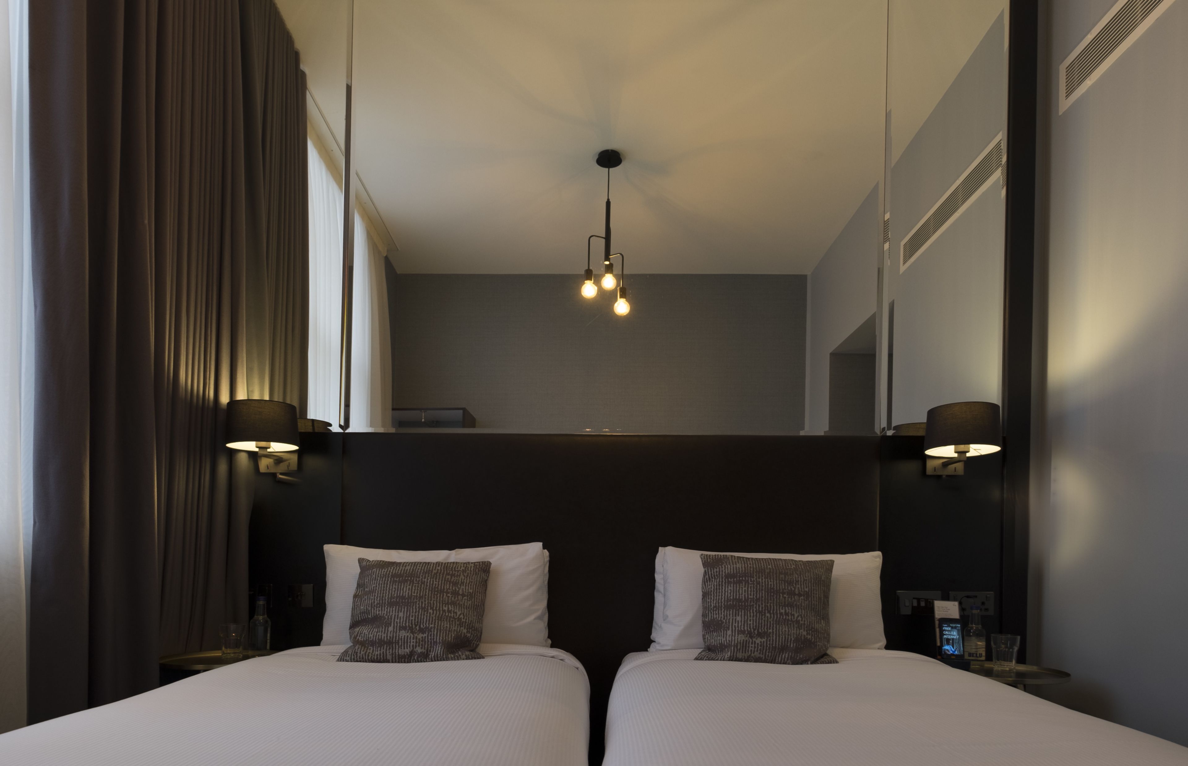 A hotel room with two beds, a ceiling light fixture, and wall-mounted lamps.