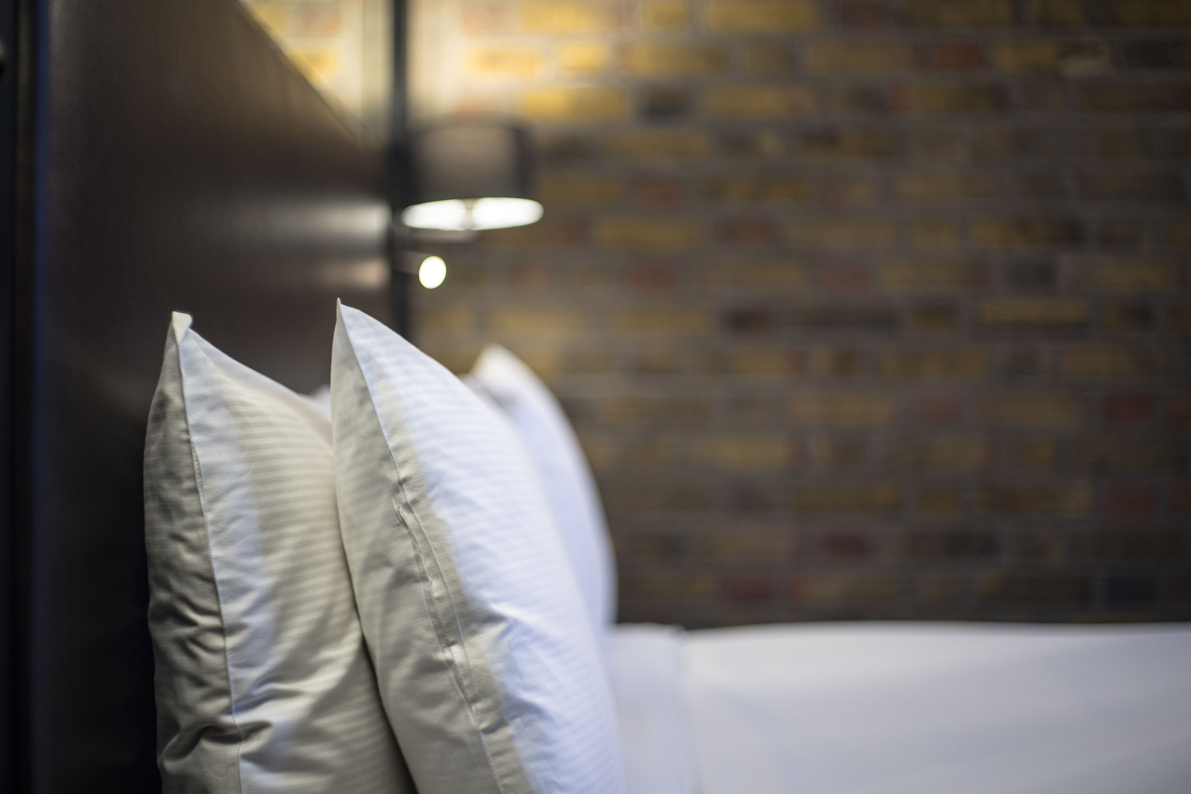 A close-up of a bed with white bedding and pillows, set against a brick wall with a modern light fixture.