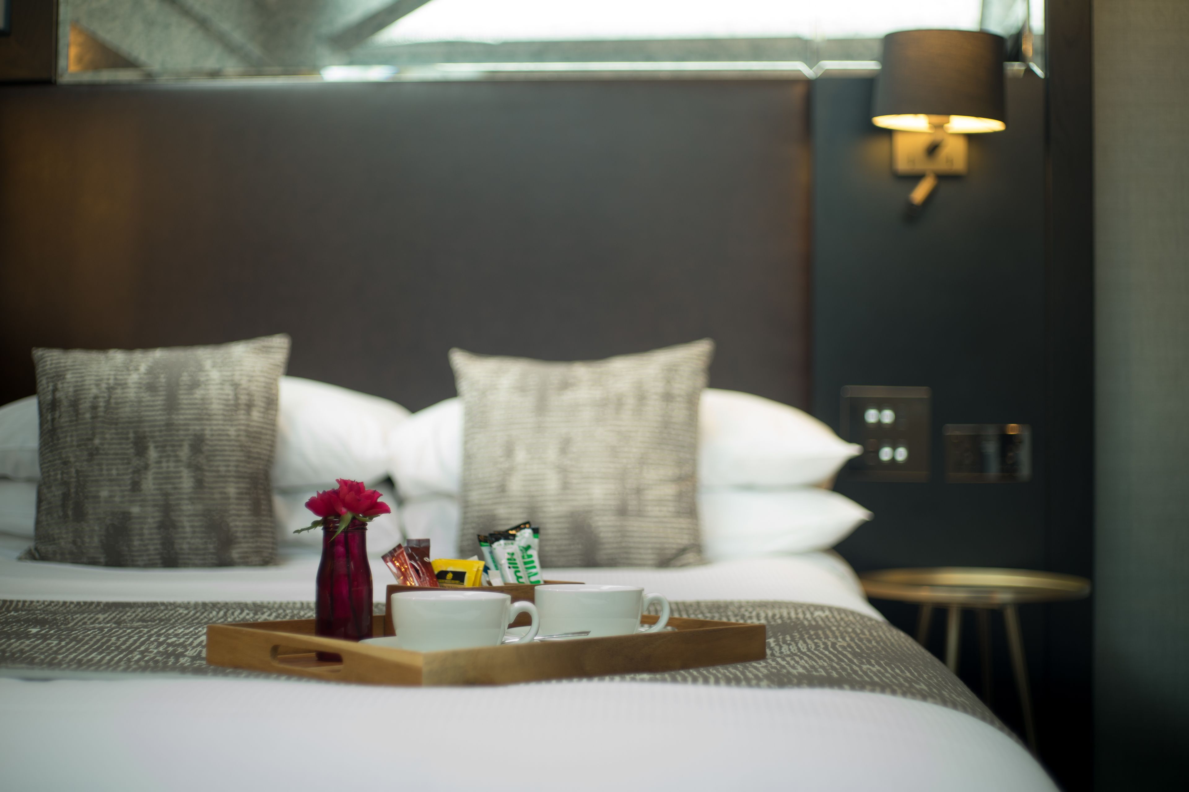 Una cama bien hecha con almohadas, una bandeja con un florero y una taza, en una habitación moderna con una lámpara de pared.