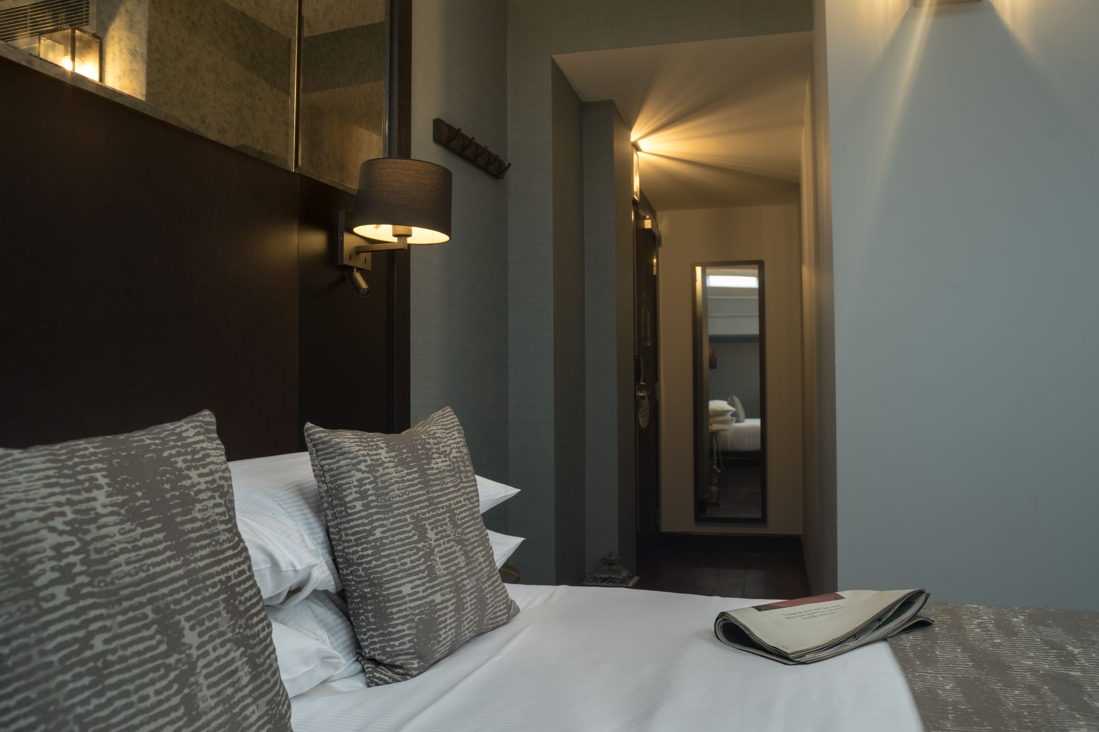 A neatly made bed with patterned pillows and a bedside lamp, a mirror on the wall, and a hallway leading to a room with a bed.