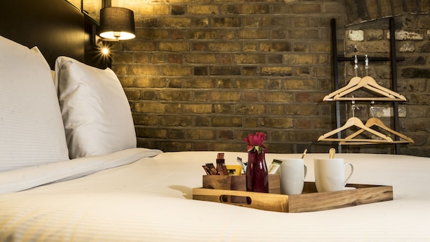 A neatly made bed with pillows, a tray with a vase and cups, and a brick wall with a hanging rack. - Camden Enterprise Hotel & Pub (London)