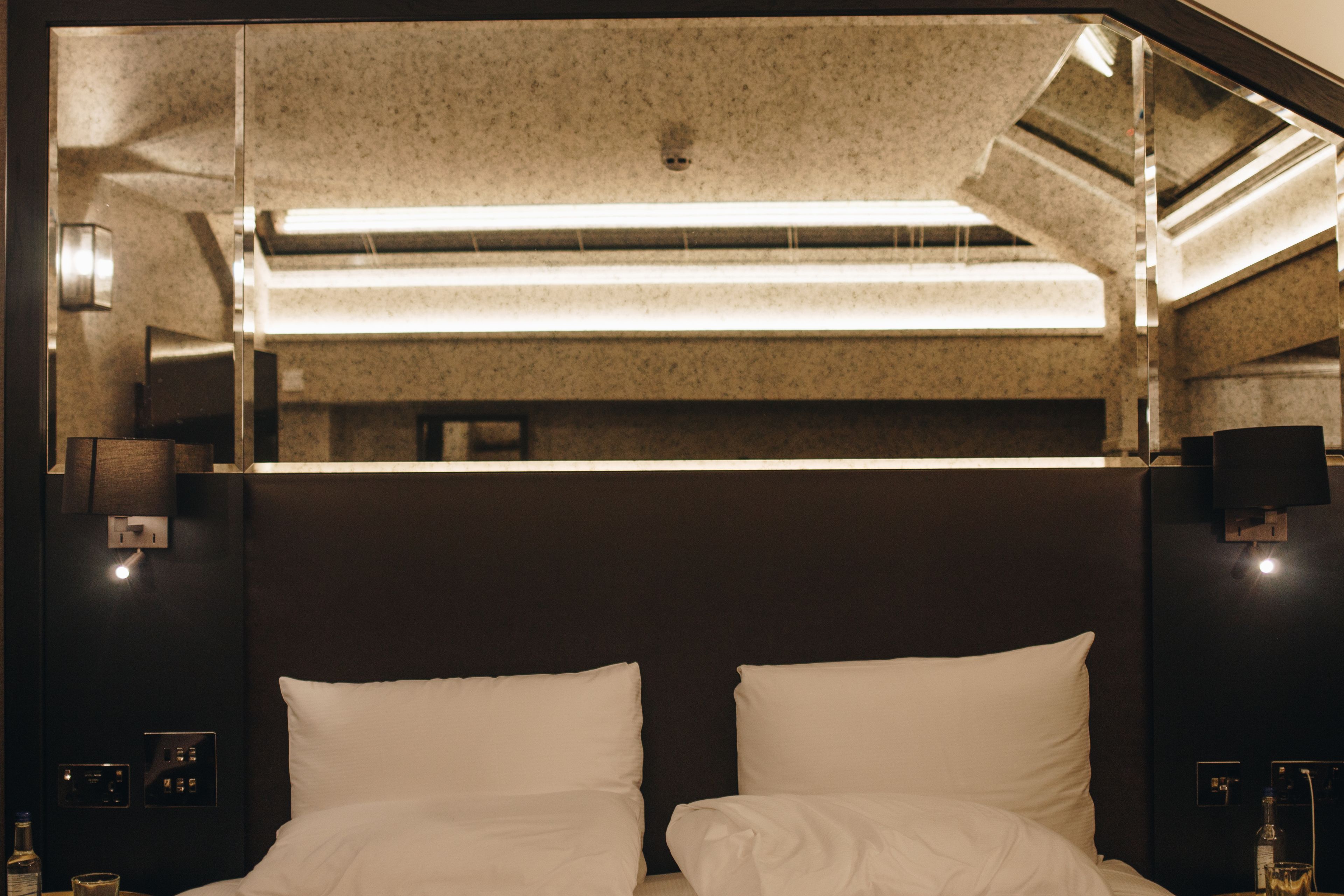 Une chambre d’hôtel avec un grand lit, une tête de lit sombre et un mur miroir qui reflète les lumières du plafond.