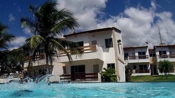 A swimming pool with clear blue water, surrounded by palm trees and a two-story building with balconies. - Apartamento Porto das Dunas - SBHS (Aquiraz)