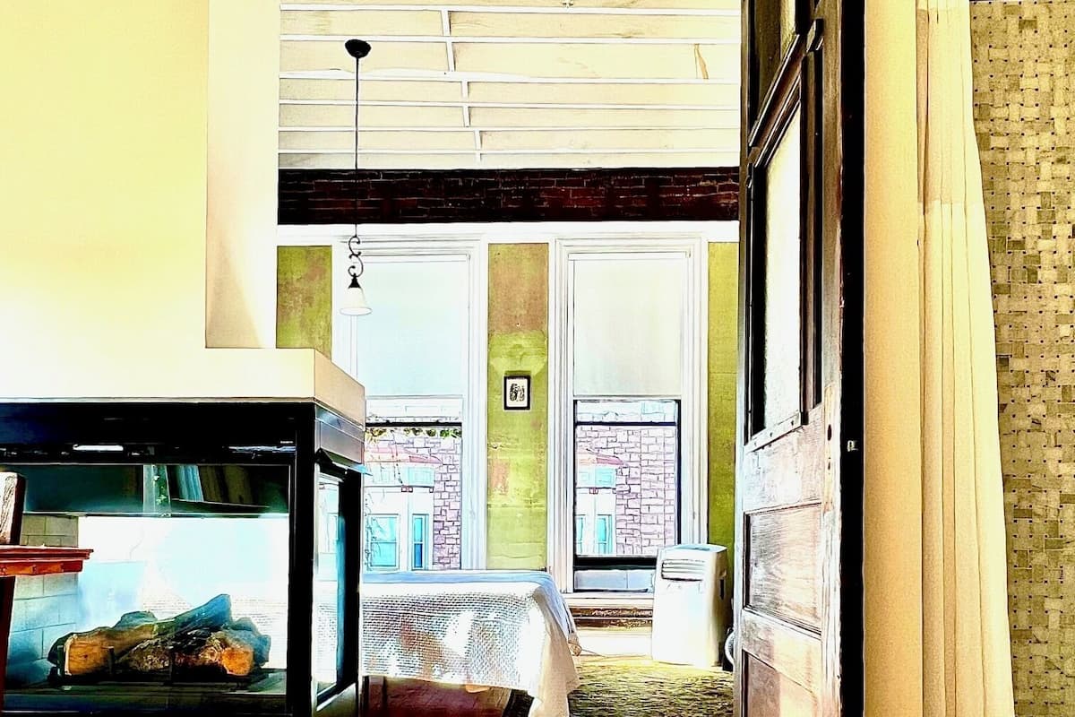 A room with a wooden floor, a bed, a fireplace, and a large window.