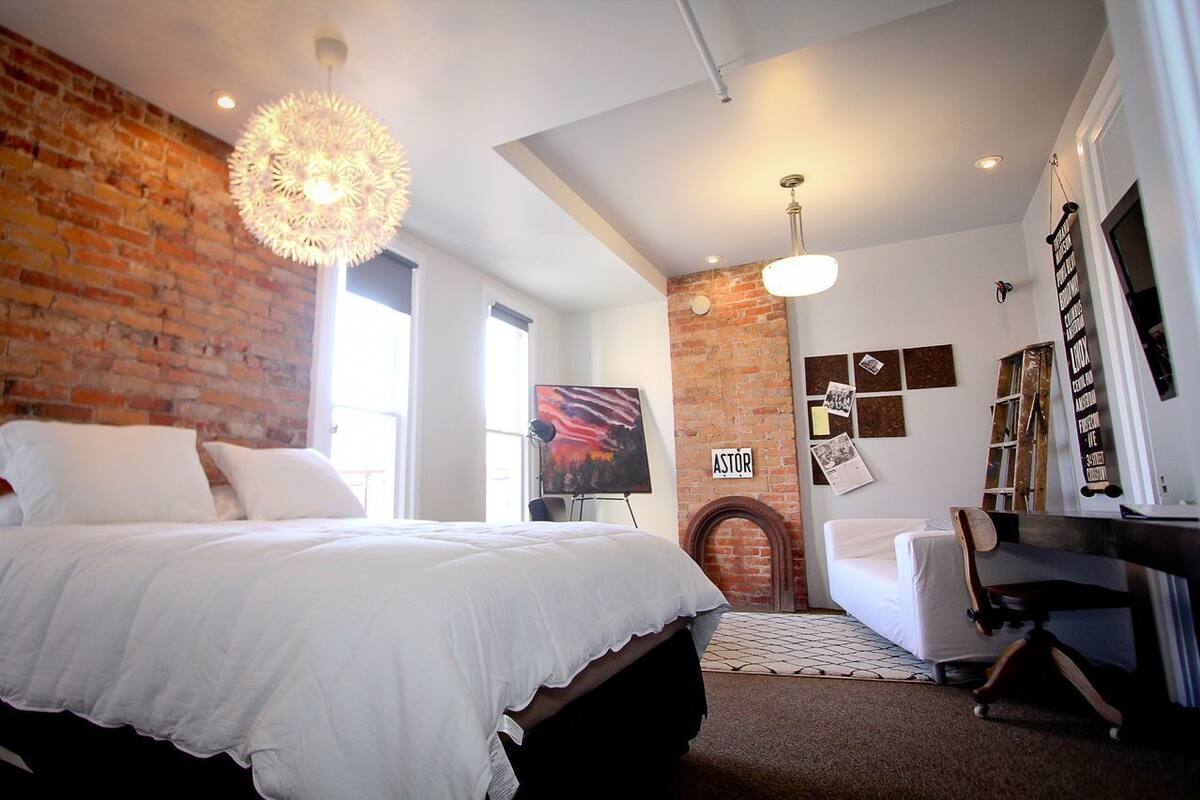 A bedroom with a brick wall, a bed with white bedding, a fireplace, and artwork on the walls.