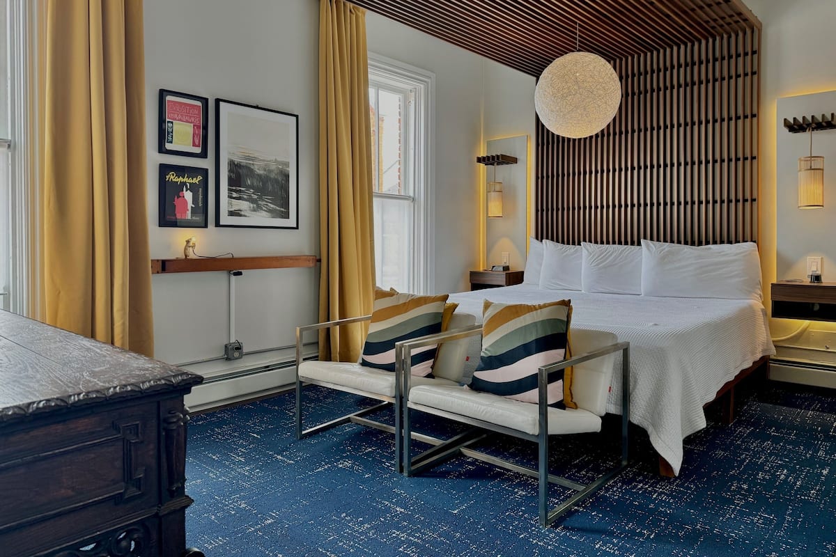 A hotel room with a bed, bedside tables, a desk, a chair, a large window, and a wall with a wooden slat design.