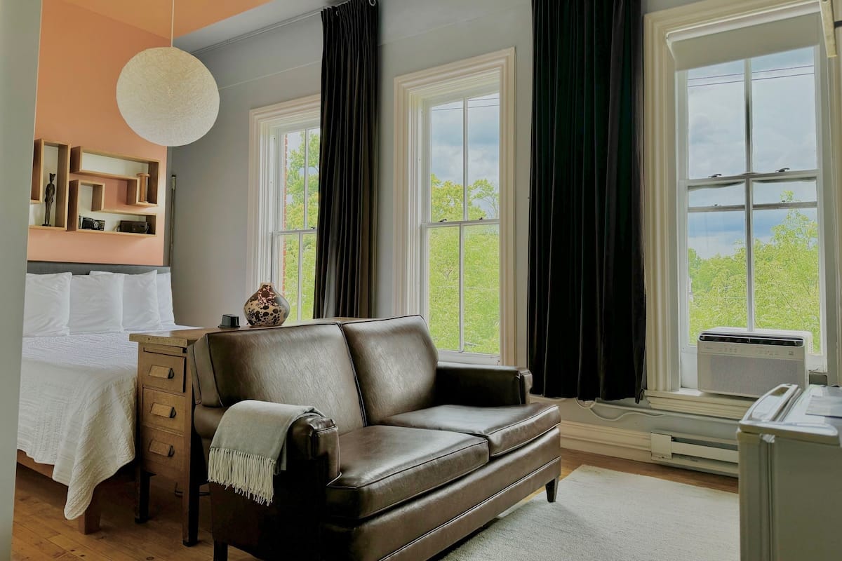 A modern room with a leather sofa, a bed with white linens, a wooden nightstand, and a large window with curtains.