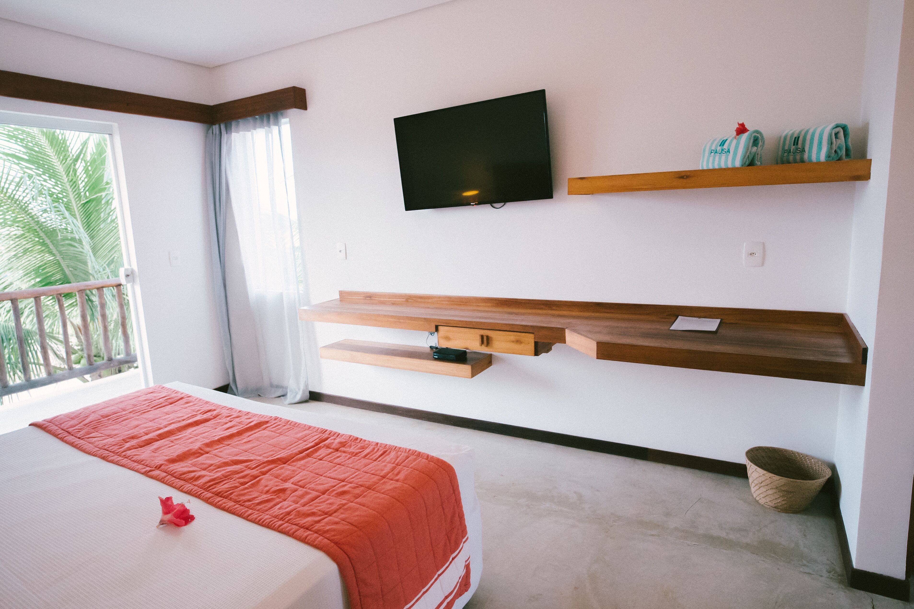 A hotel room with a bed, a TV mounted on the wall, wooden shelves, a window with curtains, and a balcony.