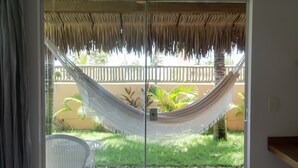 A hammock in a room with a glass door leading to a balcony with a thatched roof and a view of a garden. - Pausa Hotel (Fortim)