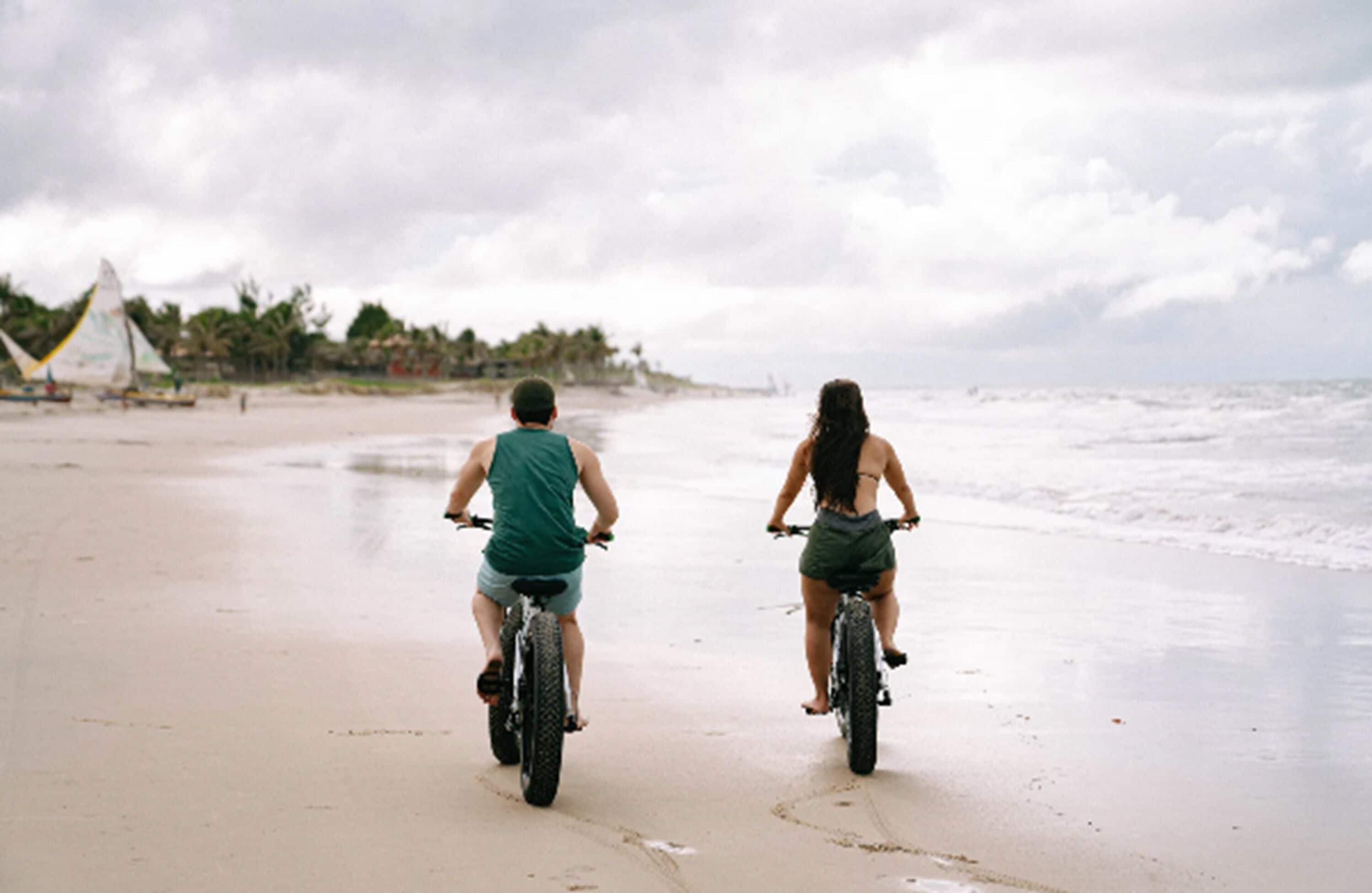 Dos personas paseando en bicicleta sobre una playa arenosa, con cielo nublado y una vista distante de un velero y palmeras.