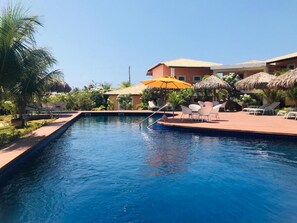Une piscine entourée de chaises longues et de parasols, bordée de palmiers et d’un bâtiment doté d’un toit en tuiles.