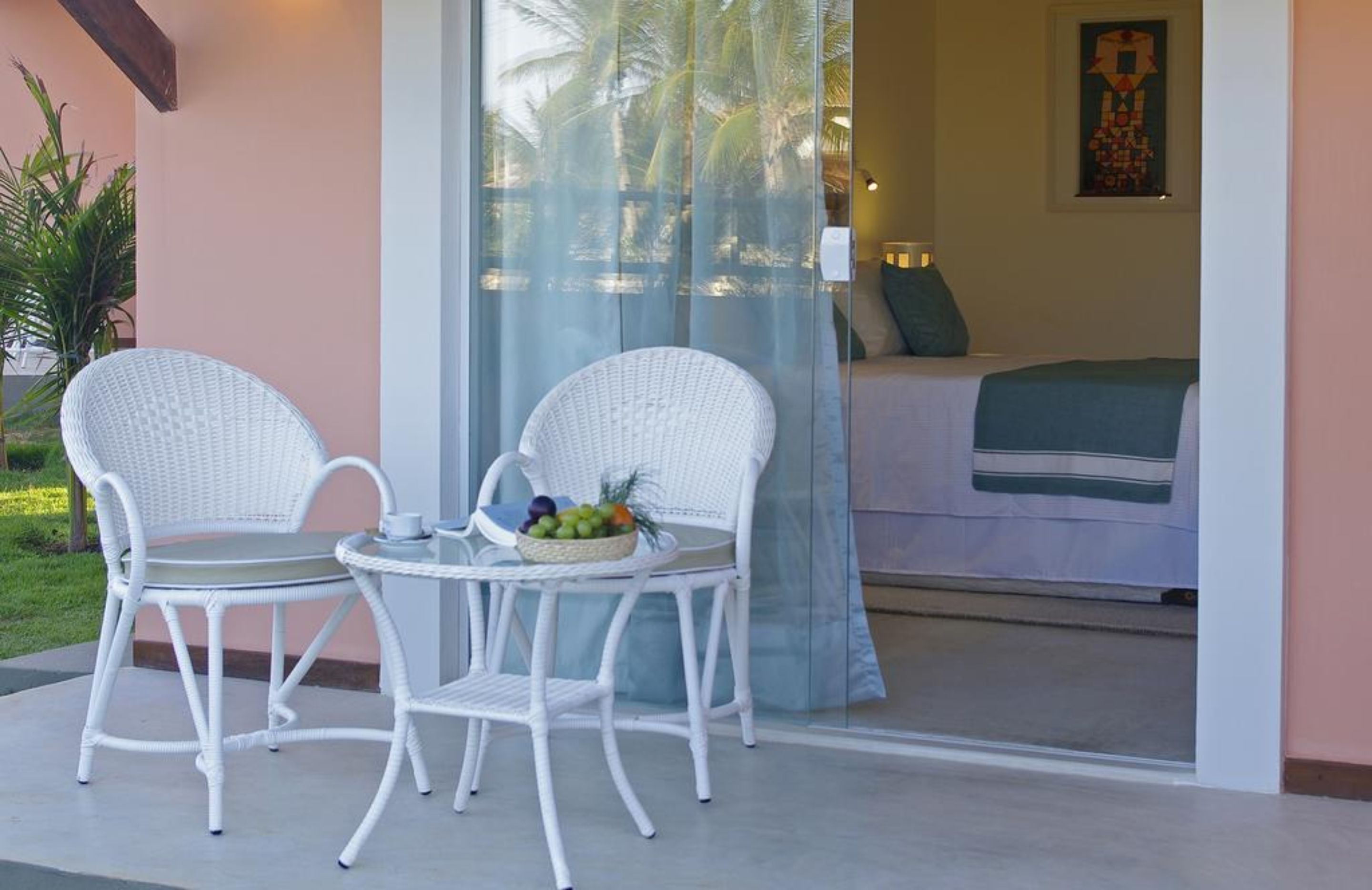 A room with a bed, a glass door leading to a balcony with two wicker chairs and a table with fruit.