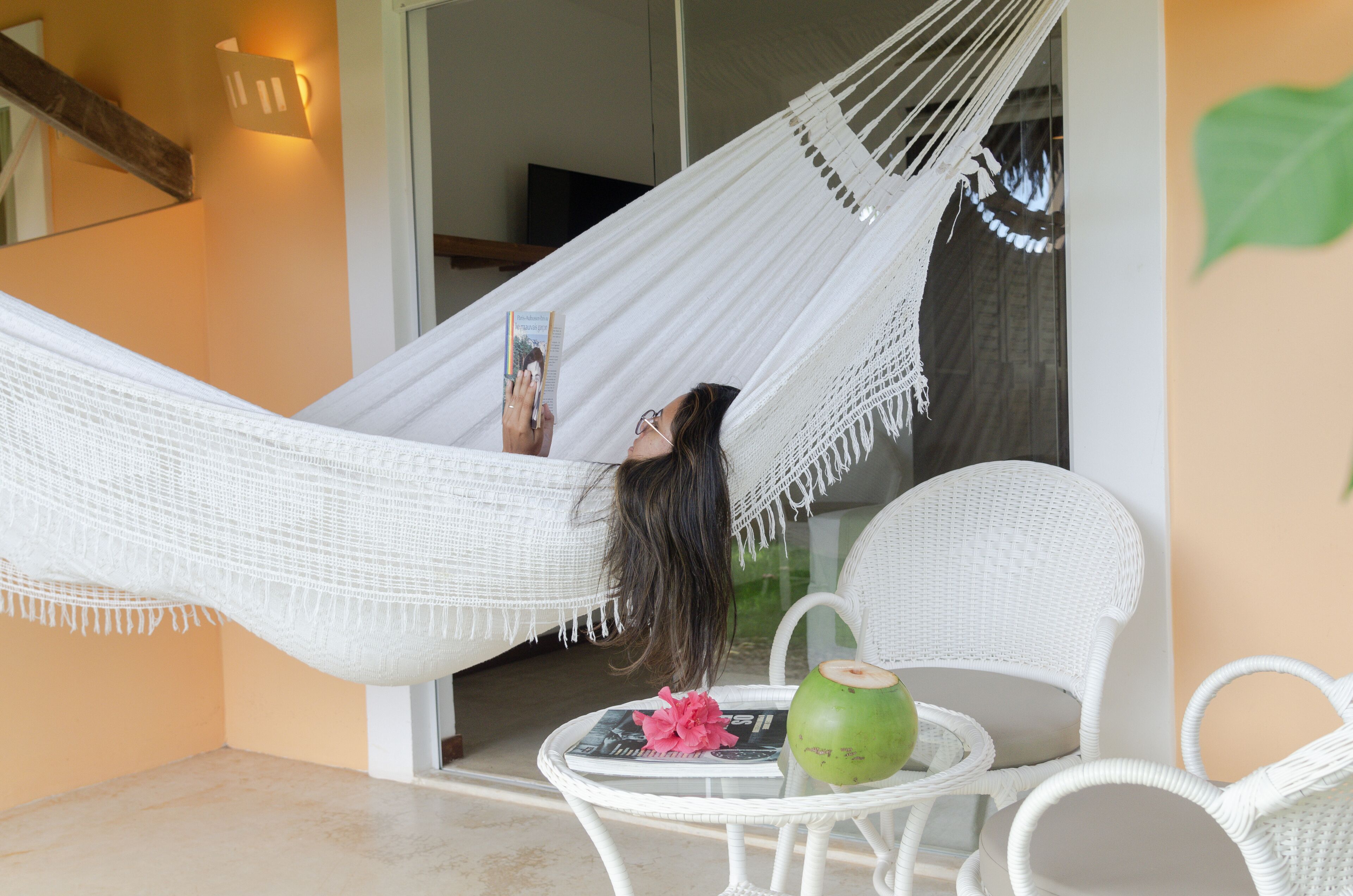 A hammock chair with a book, a green coconut, and a pink flower on a table.