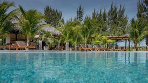 Un área junto a la piscina con sillones de descanso, palmeras y una estructura con techo de paja.
