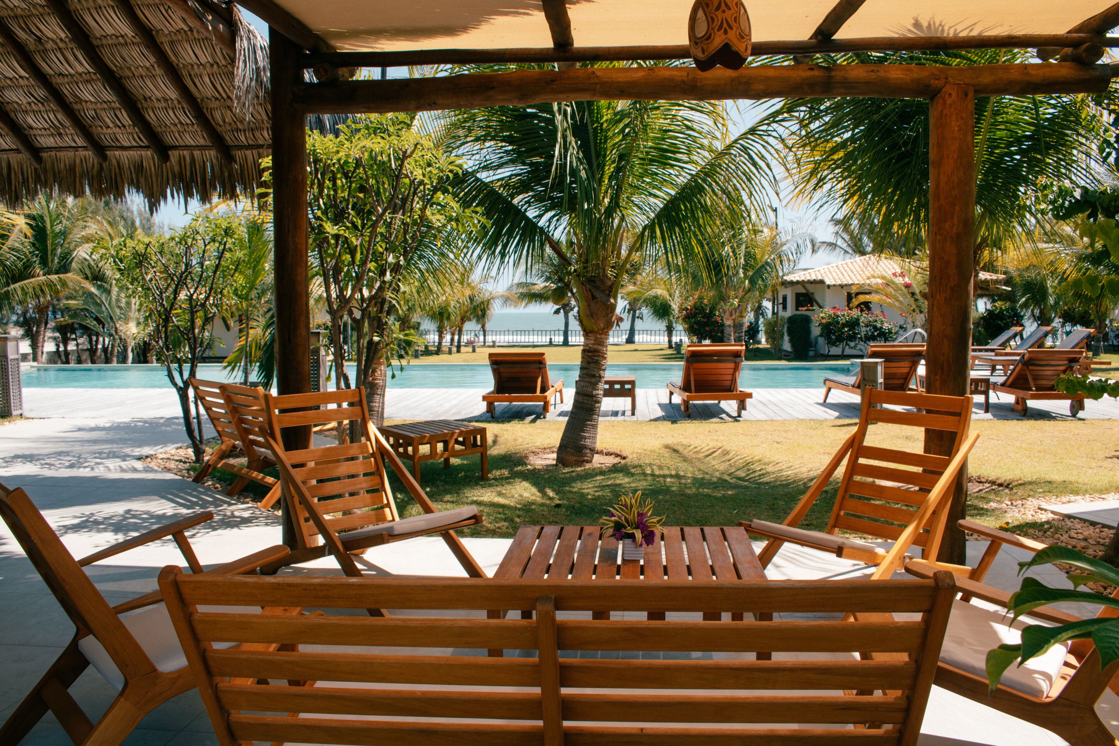 Terraza de madera con sillones de descanso y una mesa, con vista a una piscina y palmeras.