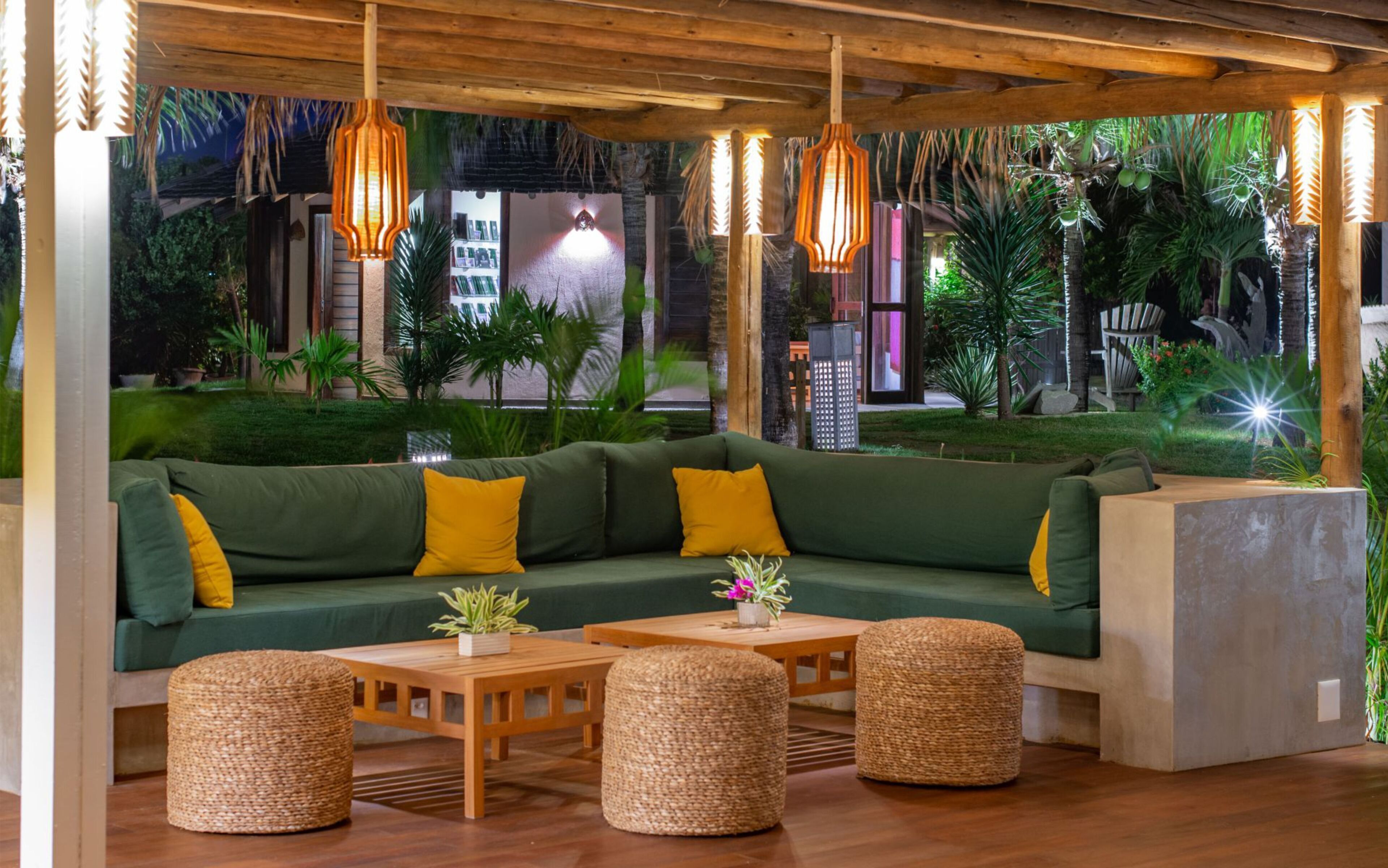 A cozy lounge area with a green sofa, yellow cushions, and woven ottomans. There are hanging pendant lights and a view of lush greenery outside.