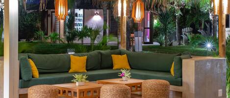 A cozy lounge area with a green sofa, yellow cushions, and woven ottomans. There are hanging pendant lights and a view of lush greenery outside.