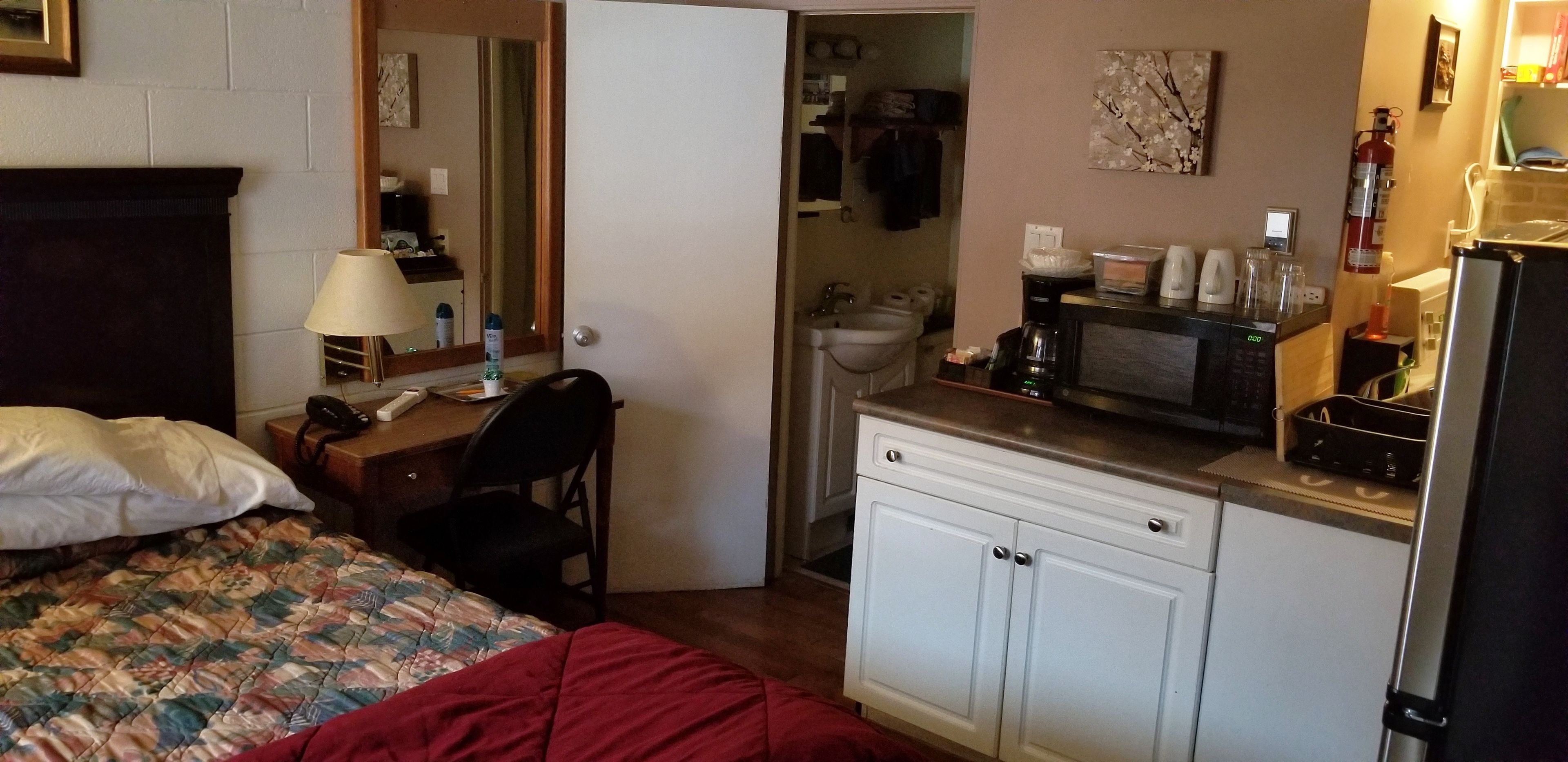 Habitación de hotel compacta con cama, escritorio, silla, microondas, refrigerador y una pequeña kitchenette.