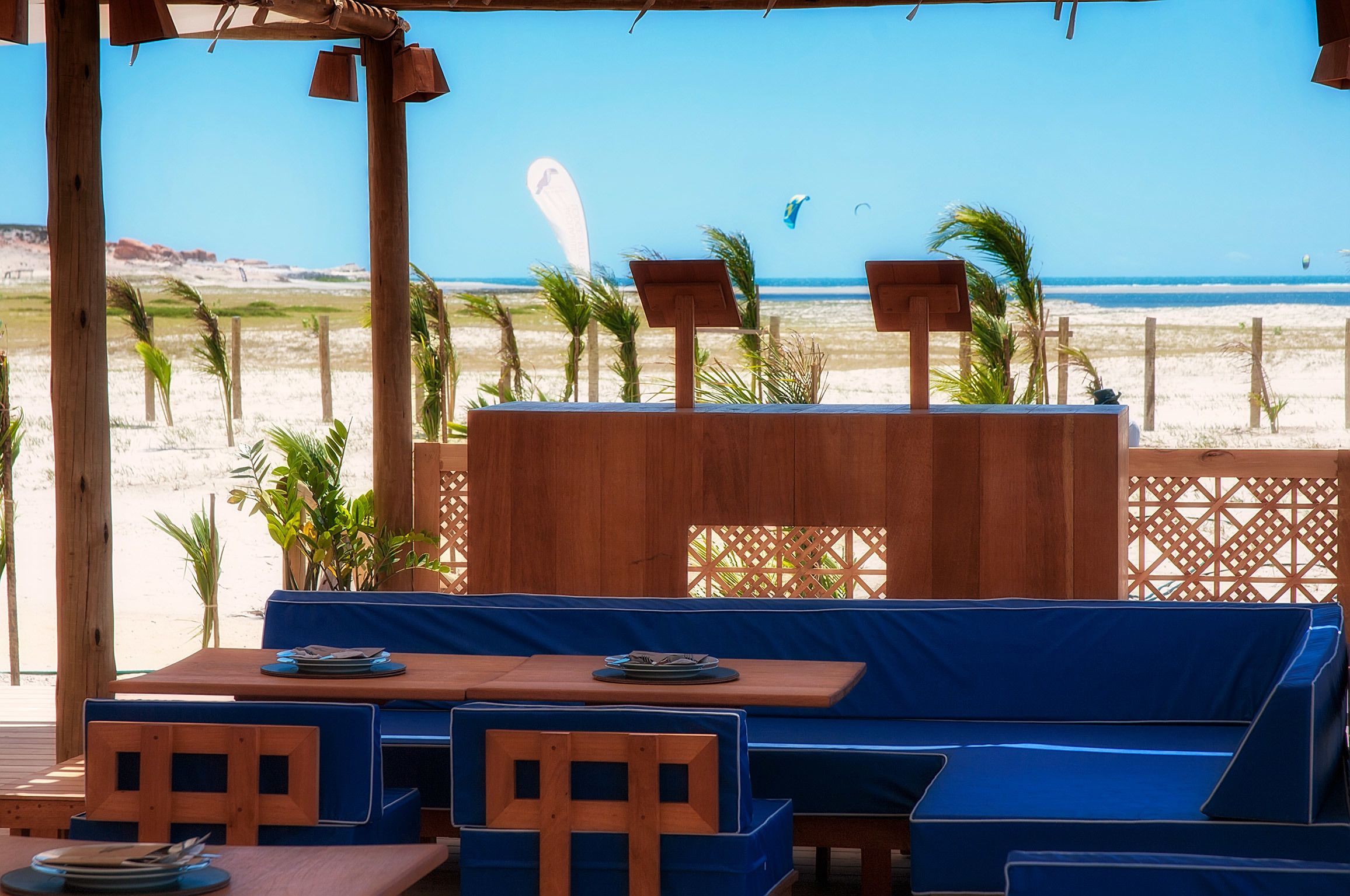 Un área de descanso junto a la playa con mesas de madera y bancos acolchados azules, con vista al océano.