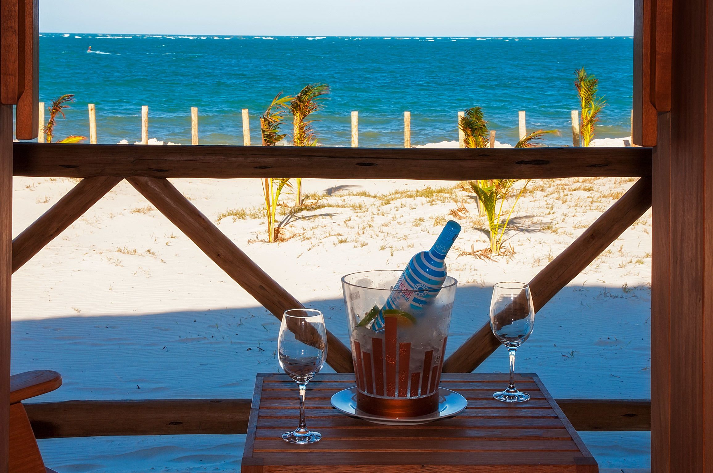 A wooden deck with a table set for two, overlooking a beach with a clear view of the ocean.