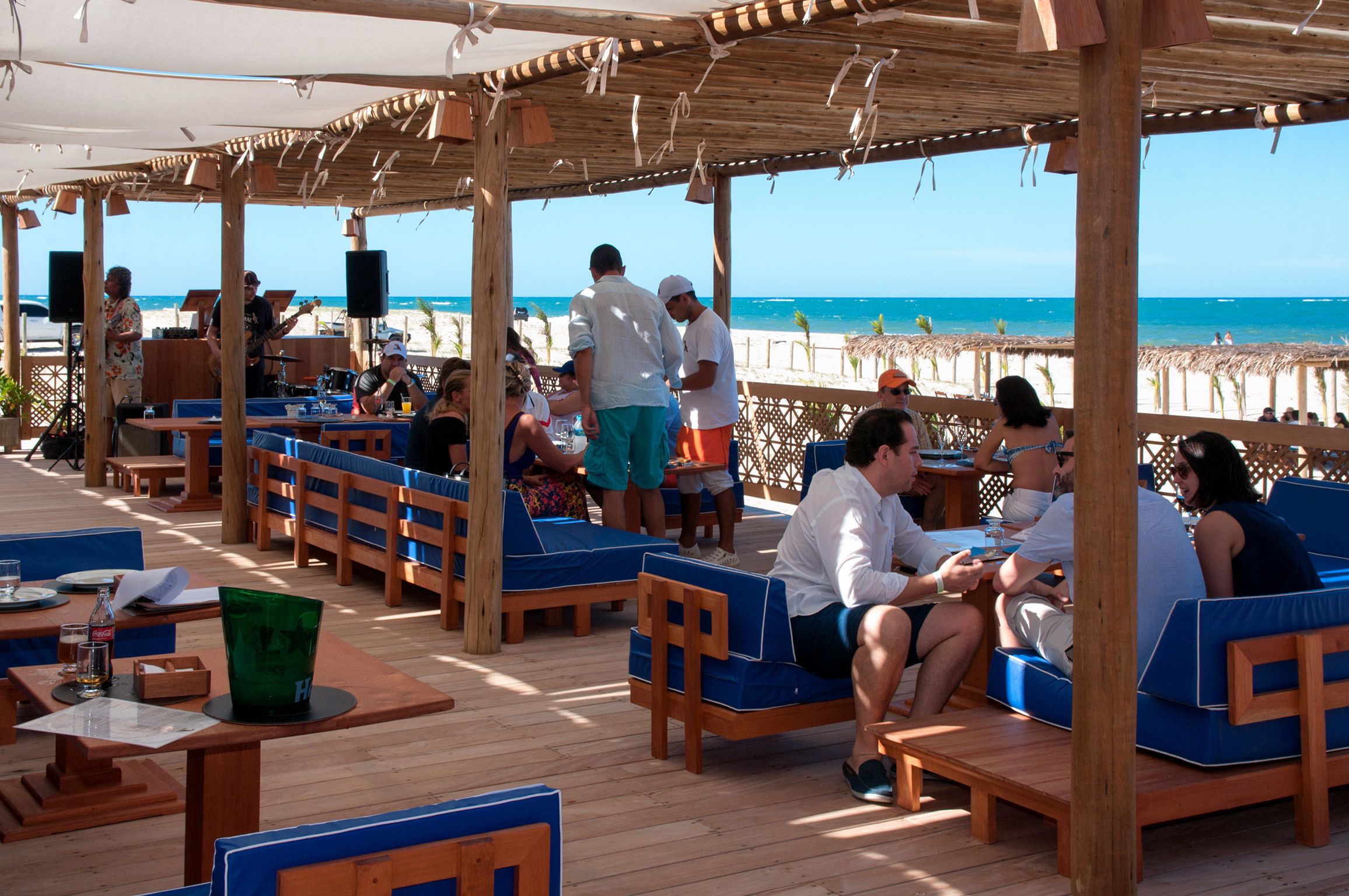 An outdoor seating area with wooden tables and blue cushioned chairs, overlooking the beach.