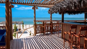 Une terrasse en bois surmontée d’un toit de chaume, offrant une vue sur une plage et l’océan.