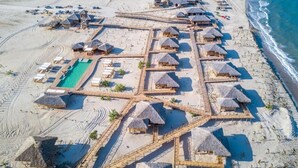 Vue aérienne d’un complexe hôtelier de bord de mer, avec des bungalows aux toits de chaume disposés selon un plan en forme de grille, une piscine centrale et une plage de sable.