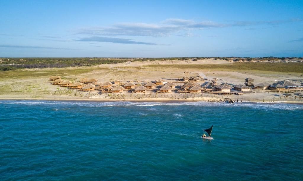 An aerial view of a coastal resort with multiple bungalow-style buildings, a sandy beach, and a clear blue sea.