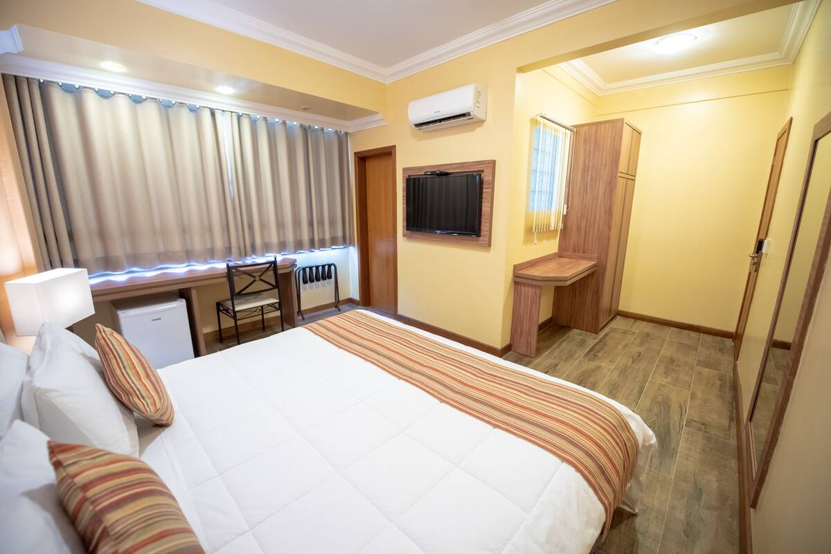 A hotel room with a bed, a desk, a chair, a TV, and an air conditioning unit.
