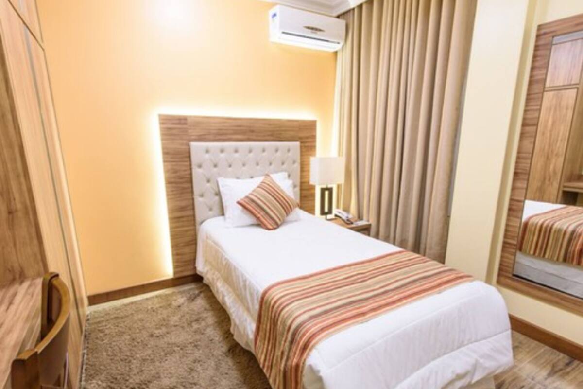 A hotel room with a bed, a nightstand, a lamp, an air conditioning unit, and a mirror.