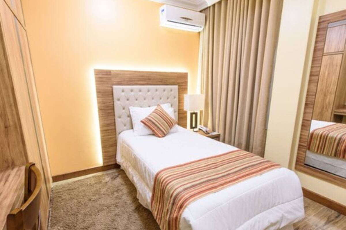 A hotel room with a large bed, a headboard, a bedside table with a lamp, a mirror, and a window with curtains.