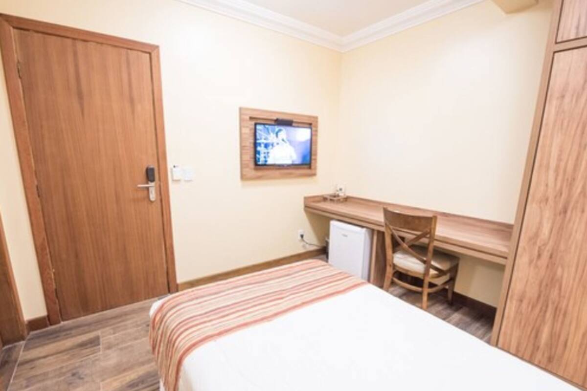 A small, neatly arranged room with a bed, a desk, a chair, and a television on the wall.