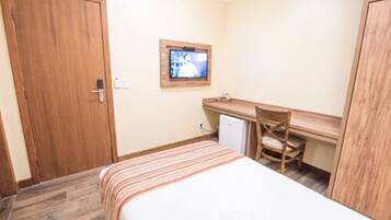 A small, neatly arranged room with a bed, a desk, a chair, and a television on the wall.
