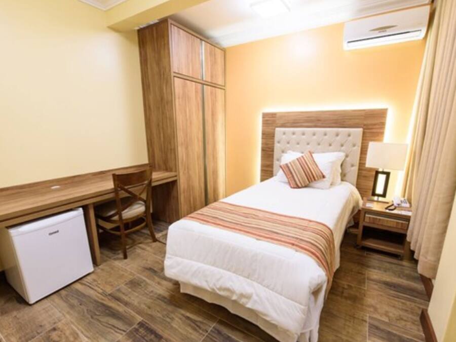 A hotel room with a bed, a desk, a chair, a wardrobe, and a small refrigerator.