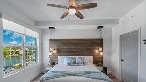 A bedroom with a large window, a ceiling fan, a bed with a wooden headboard, and bedside lamps.