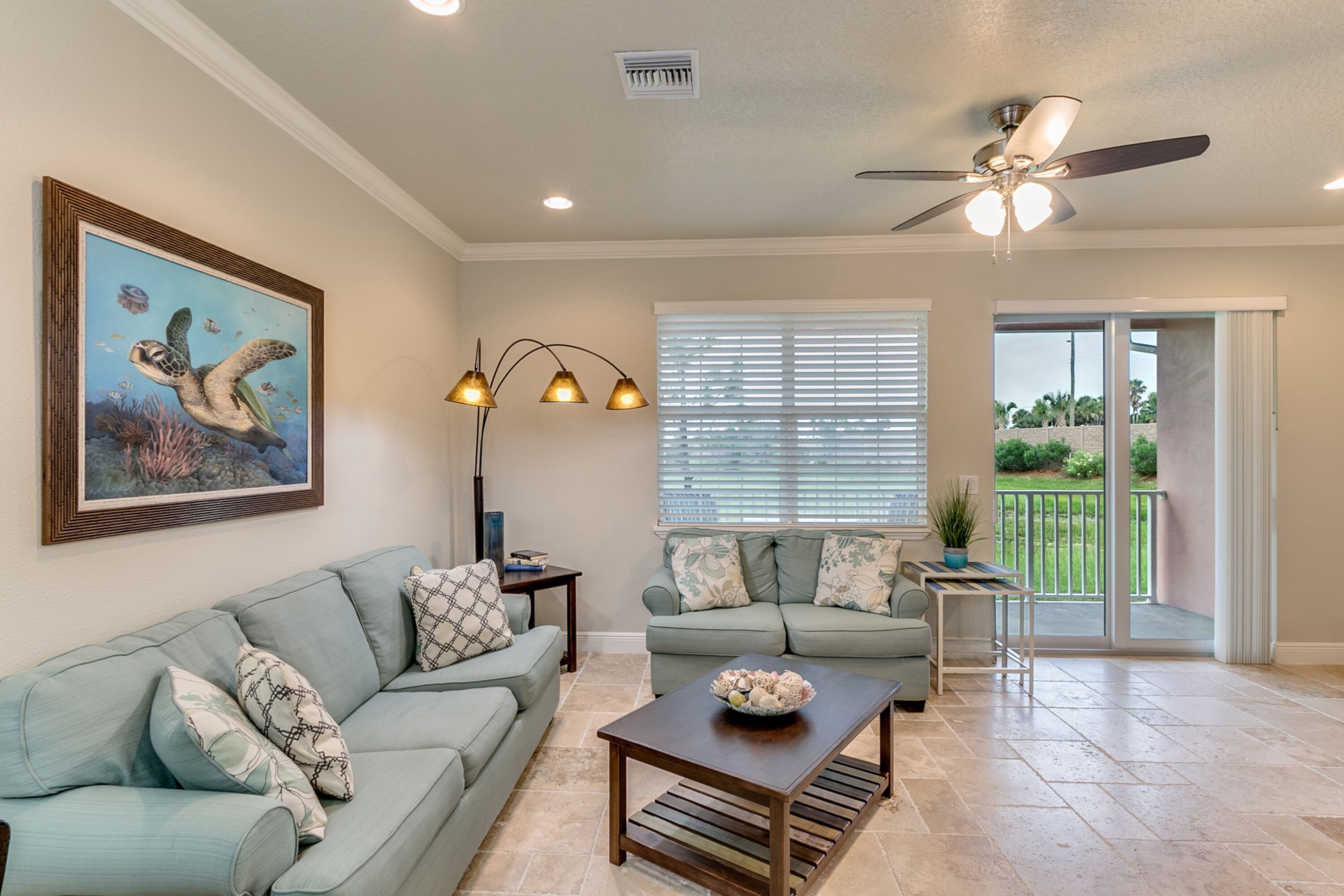 A spacious living room with a ceiling fan, a large window with blinds, a dining area with a table and chairs, and a painting of a sea turtle on the wall.