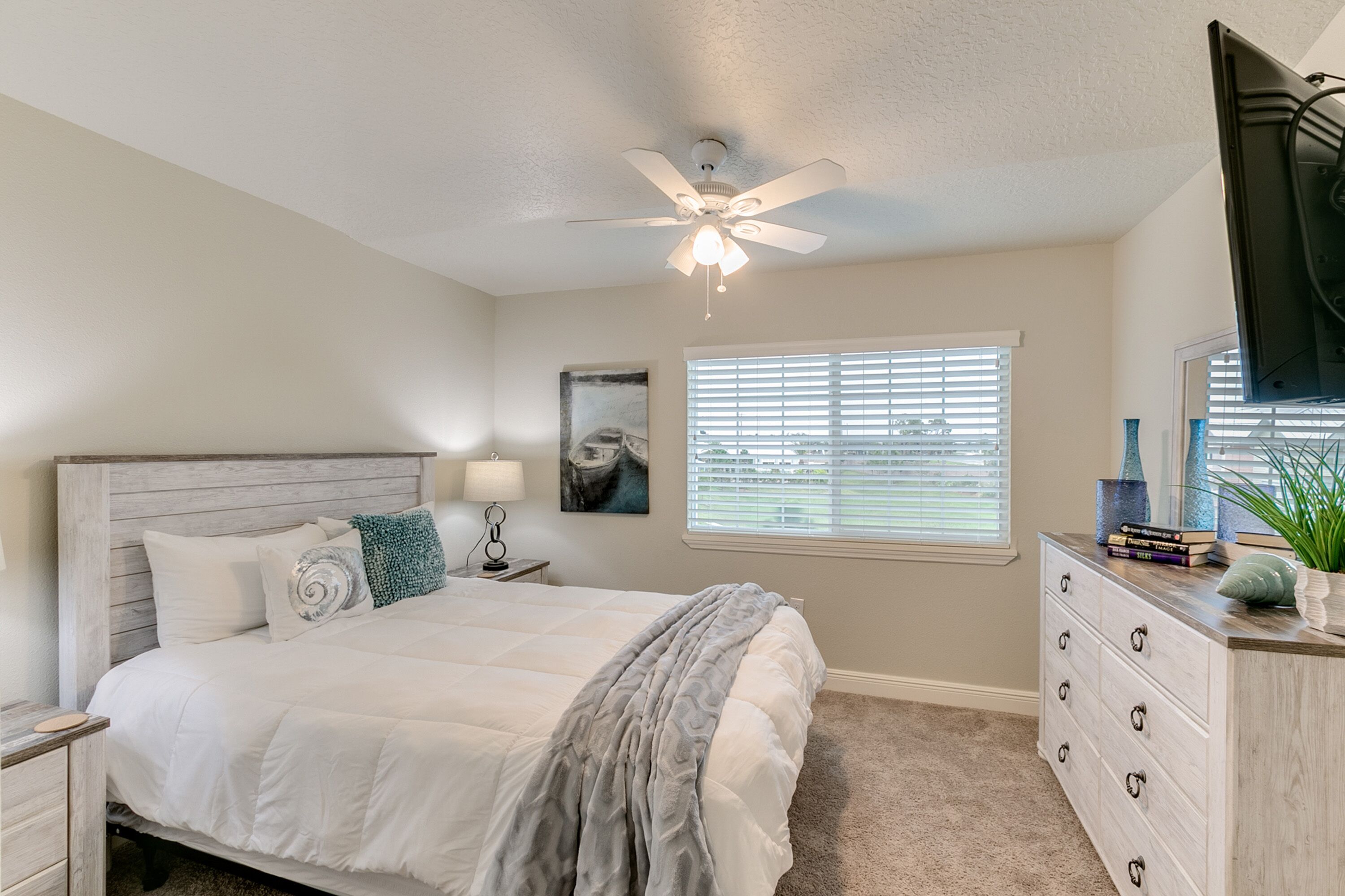 Une chambre à coucher comprenant un lit, une table de chevet, une commode, un ventilateur de plafond, une fenêtre avec des stores et un tableau accroché au mur.