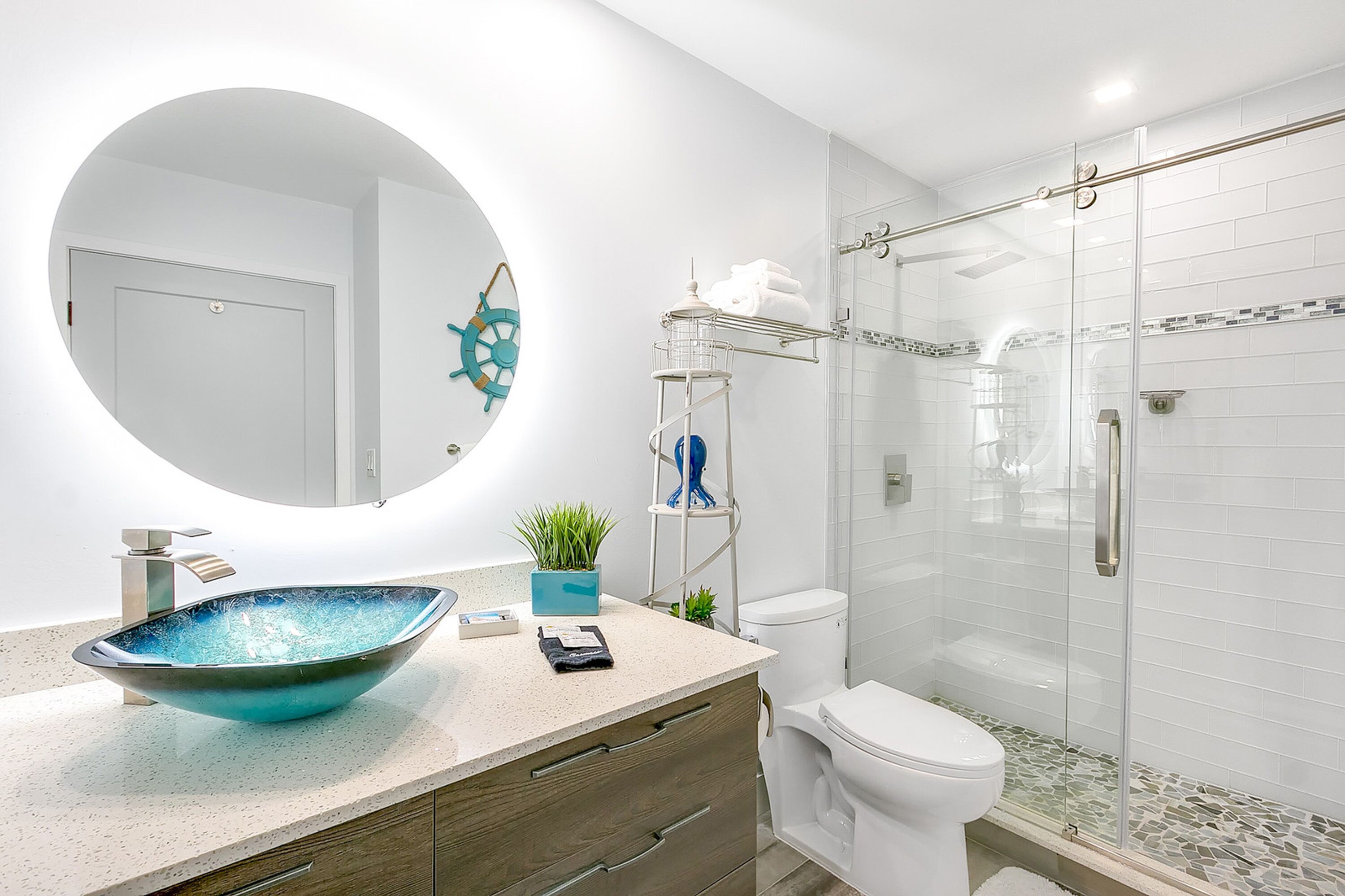 A modern bathroom with a large round mirror, a vessel sink, a toilet, and a glass-enclosed shower area.