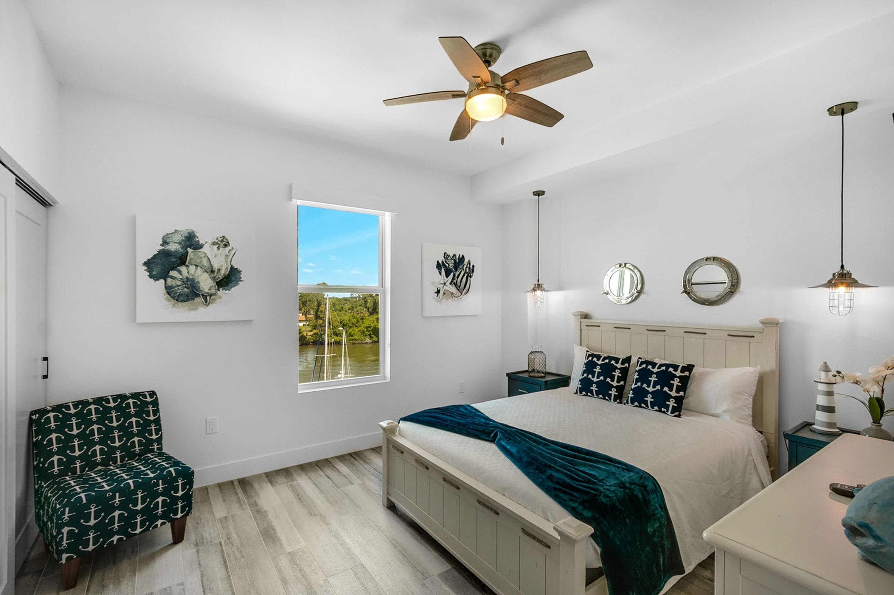 A bedroom with a large bed, a ceiling fan, a window with a view, and artwork on the walls.